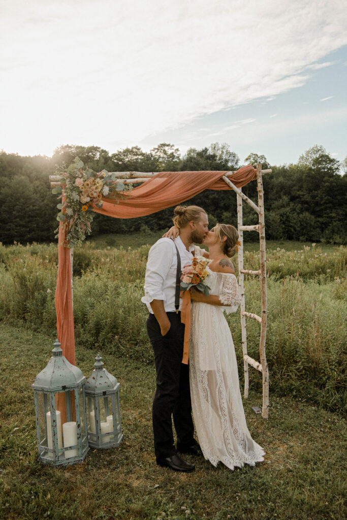 A blond bride and groom kissing under a rustic arbor in a meadow