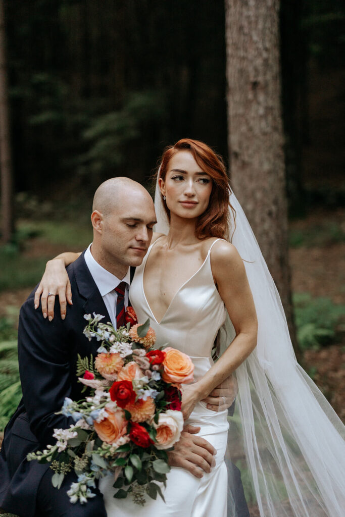 A photo by Dierdre Alston of a red-haired bride in a forest sitting on her groom's lap and holding an Idyllwild Event Design wedding bouquet