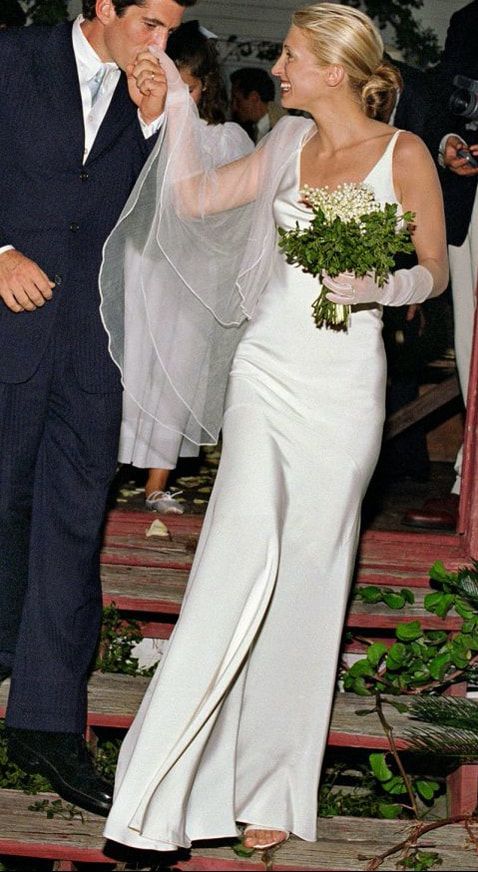 JFK Jr. kisses Caryolyn Bessette Kennedy's hand as they exit their 1996 wedding ceremony. The bride wears an impeccably fitted white satin Narciso Rodriguez bias cut gown, sheer white gloves and veil, and carries a lily-of-the-vally bouquet.