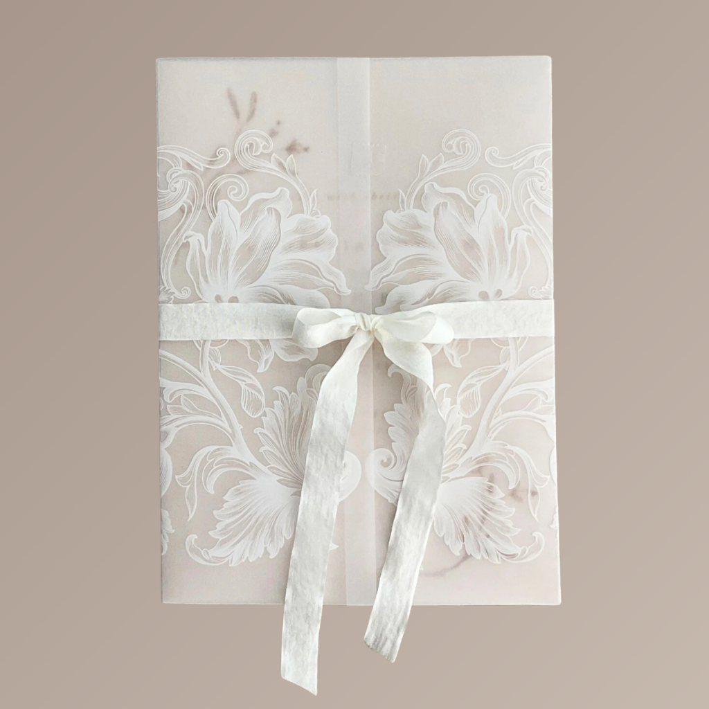 An opulent wedding invitation wrapped in white vellum with flowers printed in white ink. The classically beautiful invitation is tied with a dainty white silk bow.