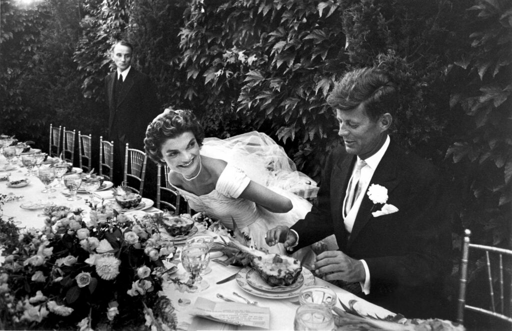 Black and white photo of Jacqueline Kennedy and John F. Kennedy at their wedding reception. Jacqueline, in a classic white gown, leans joyfully towards John, who is in a tuxedo with a boutonnière, both seated at a long, elegant table adorned with floral arrangements and crystal glassware.