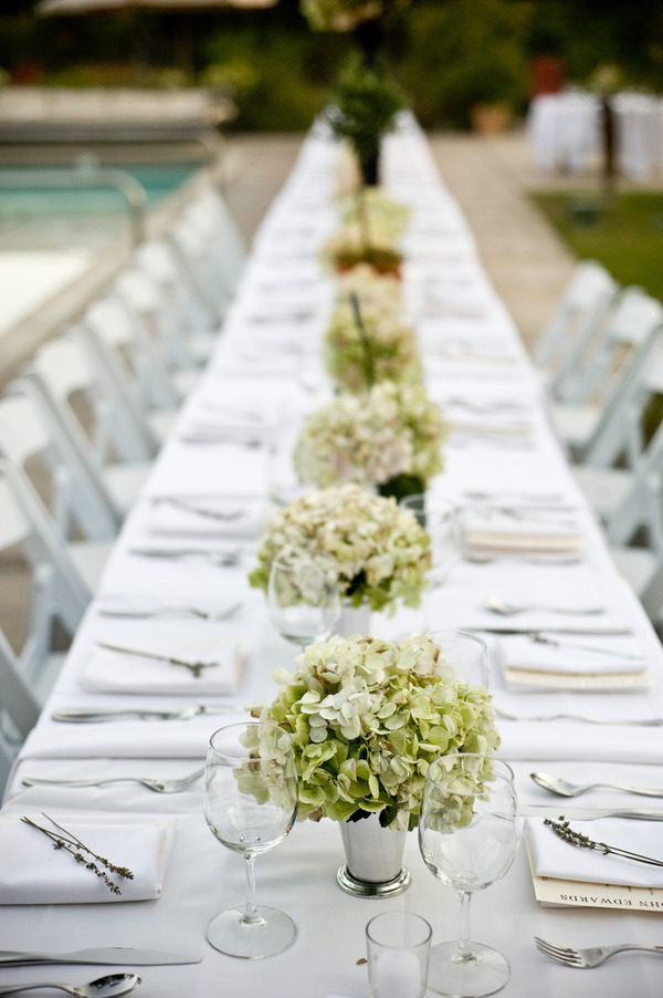 A wedding reception table dressed in white linens, featuring many rows of silver julep cups with green hydrangeas.