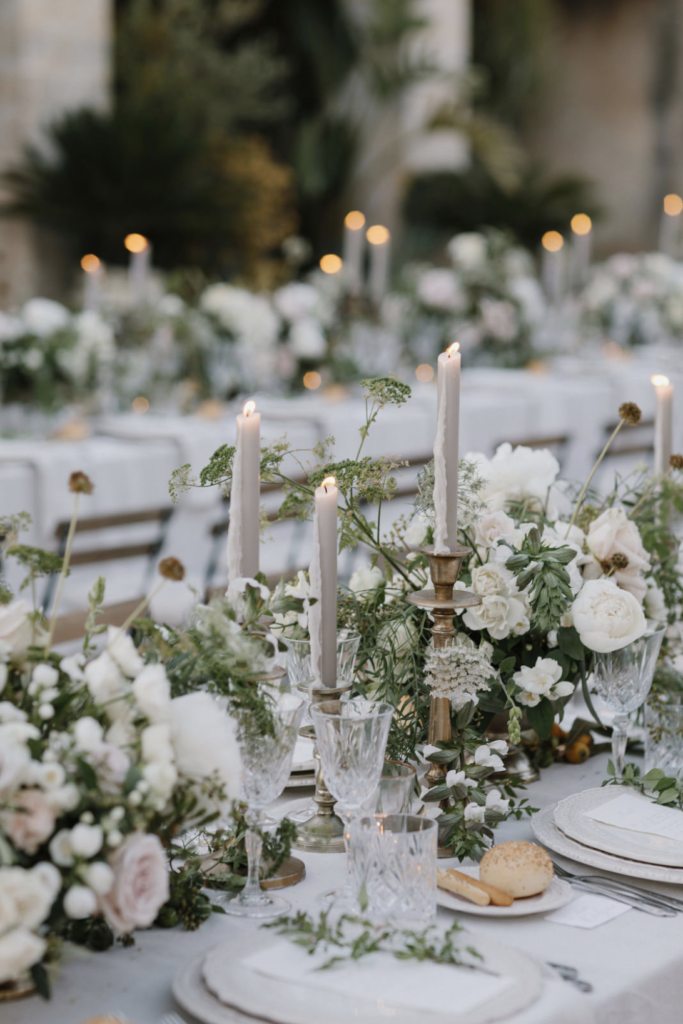 A sophisticated table setting featuring a gray monochromatic palette, garden-style centerpieces with peonies, brass tapers, and textured plates and stemware create a refined and opulent ambiance for an old money wedding.