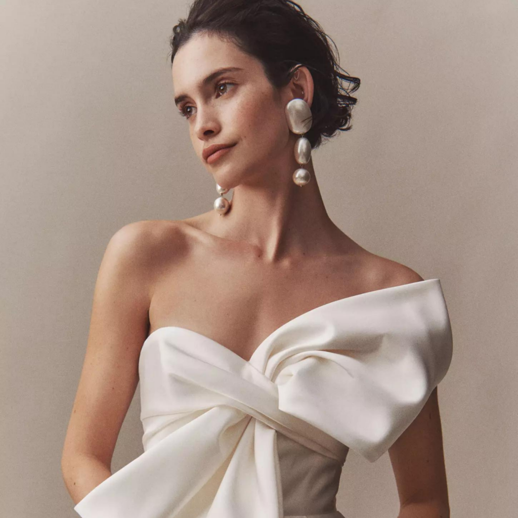 A bride models an off-the-shoulder white wedding gown with a large, stylish bow on the front. She wears statement pearl earrings and has short, dark hair styled in soft waves, exuding modern elegance against a neutral background.