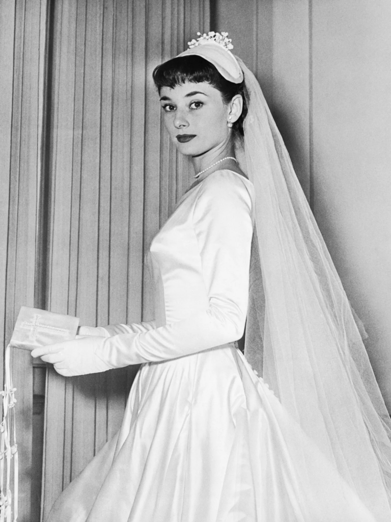 A portrait of Audrey Hepburn wearing a tailored, elegant bridal gown with simple lines and luxurious fabric, reflecting the understated luxury and timeless beauty suitable for an old money wedding.