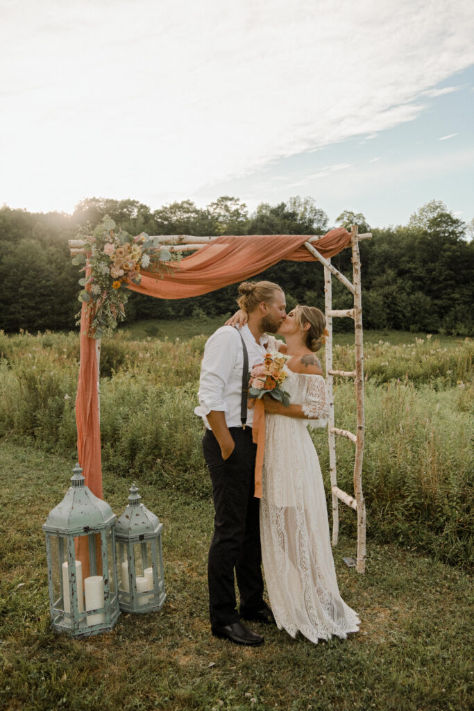 Bride and groom kiss under a floral arch adorned with warm-hued blooms, perfectly complementing the natural outdoor Catskills wedding venue