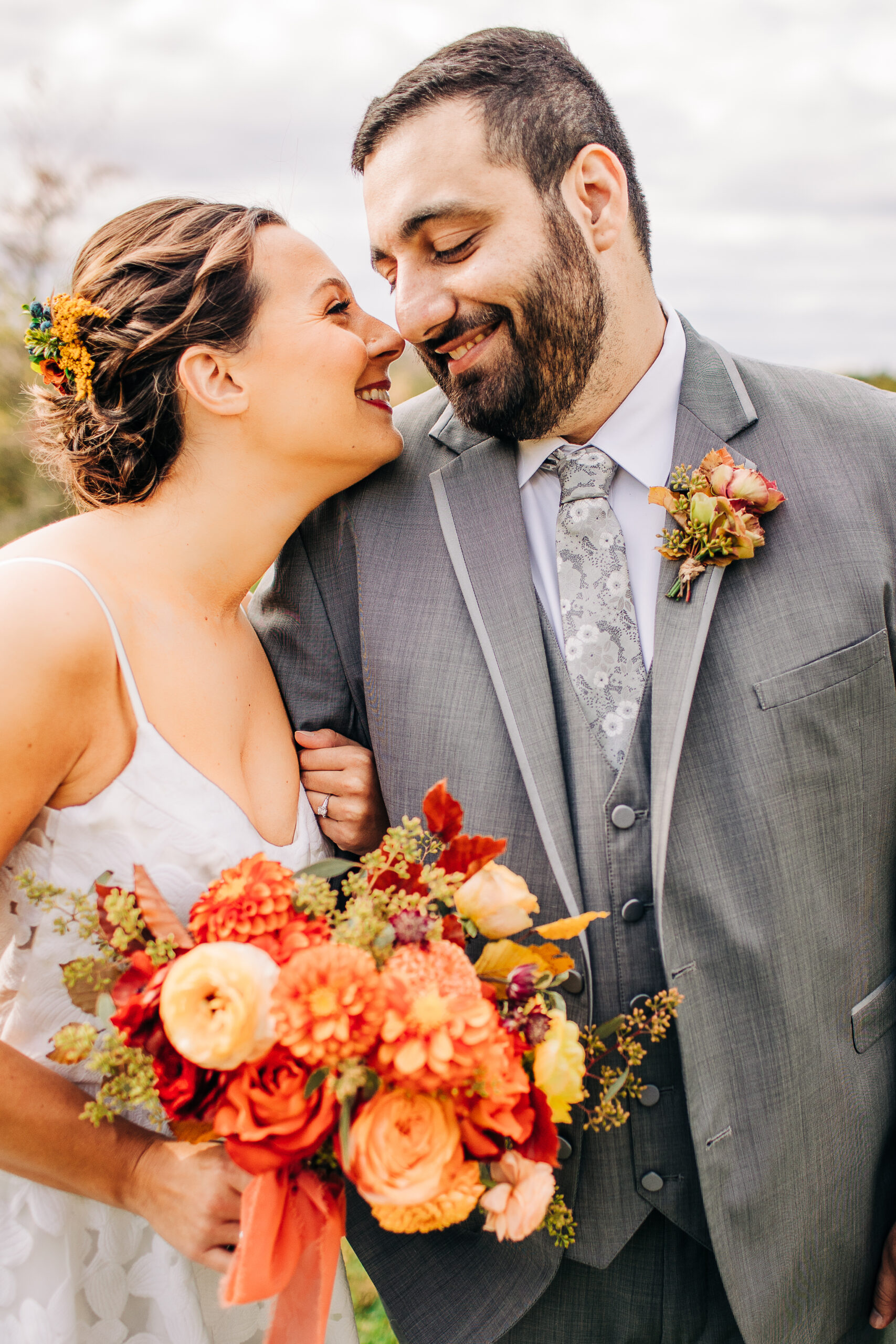 Bride with autumn dahlia and ranunculus bouquet and groom in a gray suit touch noses