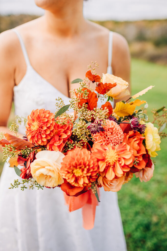 Bride holds a custom jewel-tone bouquet featuring orange dahlias and greenery, crafted by Idyllwild Event Design for a fall wedding in Washingtonville, NY