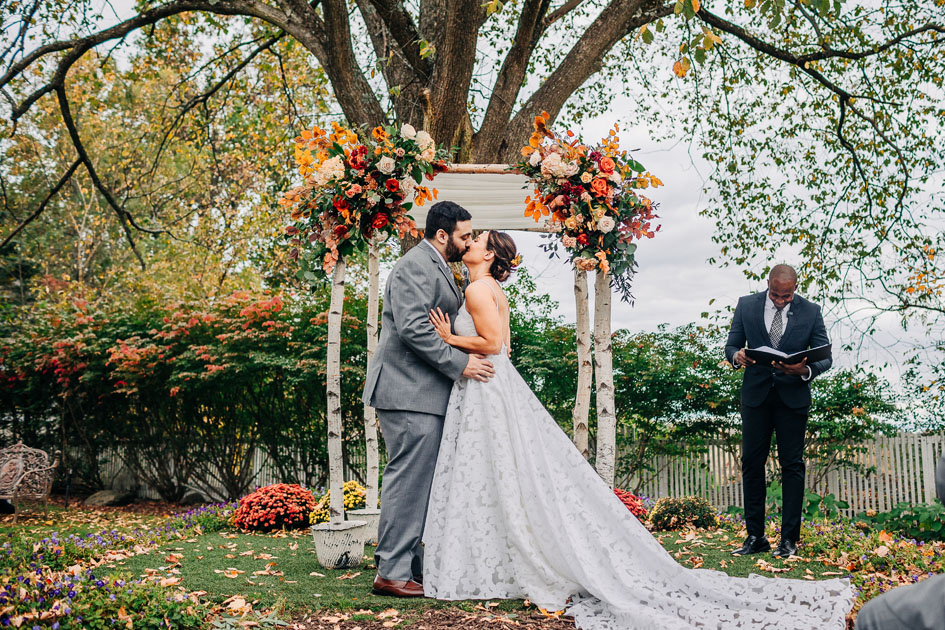 Bride and groom kiss beneath a birch chuppah decorated with vibrant fall flora and foliage at their Hudson Valley autumn wedding. Floral by Idyllwild Event Design