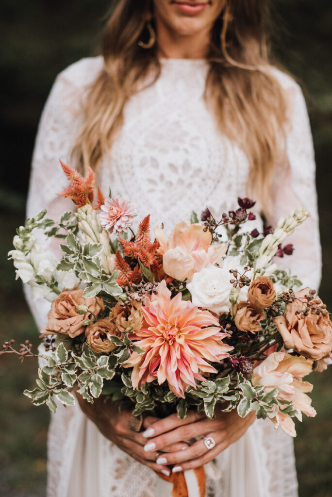 Bridal bouquet in copper, peach, and ivory tones with wild greenery and Queen Anne’s lace, held against a lace gown with sleeves