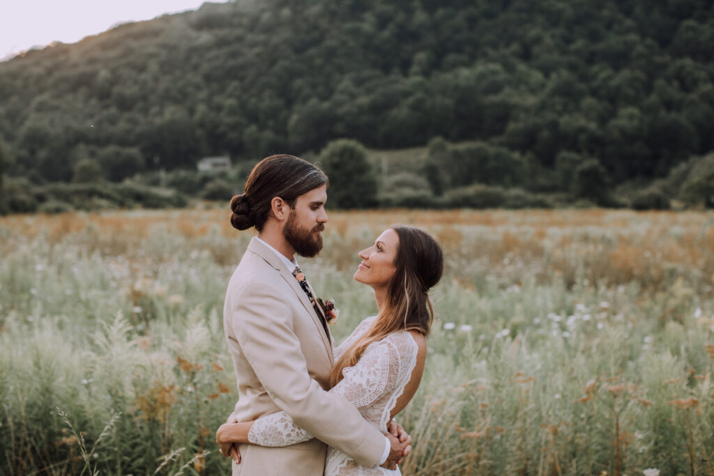 A bride and groom stand in a Catskills wildflower field with arms around one another's waists. There's a green mountain in the background.