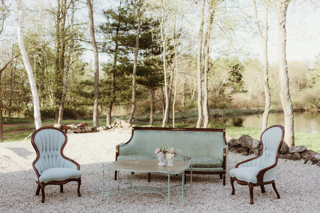 Elegant vintage wedding lounge vignette featuring a green jacquard sofa, carved wood chairs, and a pondside garden view, evoking relaxed European luxury.